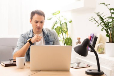 Mutlu genç adam, dizüstü bilgisayarı üzerinde çalışırken sabahın köründe kahvesini bitirmek için gülümsüyor.