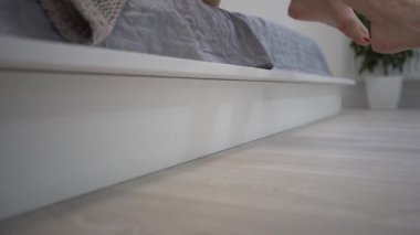 Yakın plan çekim. Bir dişinin bacakları yataktan kalkıyor. Hafif arka plan, sabah uyanır. Beyaz yataklı kadın ayak parmakları. Yere yatın. Parke ya da lamine et. fitness bacakları