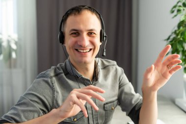 Baş ekran görüntüsü, gözlüklü ve kulaklıklı neşeli, genç yakışıklı adam kameraya bakıyor, merhaba jesti yapıyor, online video kamera görüşmesinde arkadaşlara hoş geldin diyor..