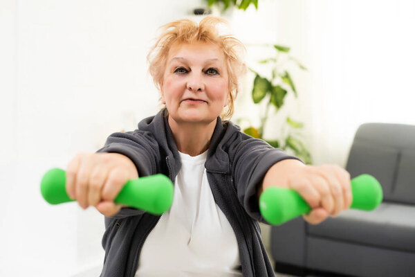 Портрет активной пожилой женщины, выполняющей гантели дома, улыбающейся.