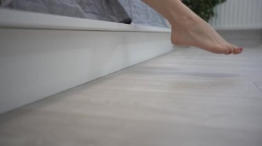 Yakın plan çekim. Bir dişinin bacakları yataktan kalkıyor. Hafif arka plan, sabah uyanır. Beyaz yataklı kadın ayak parmakları. Yere yatın. Parke ya da lamine et. fitness bacakları