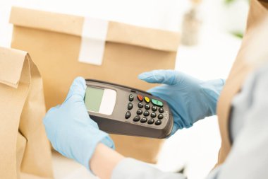 Küçük işletme, koronavirüs, sosyal mesafeler ve temassız ödeme konsepti. Kahveci, kafede garson, tıbbi maske ve eldivenler, POS terminalinin güvenli bir şekilde satın alınmasını tavsiye ediyor.