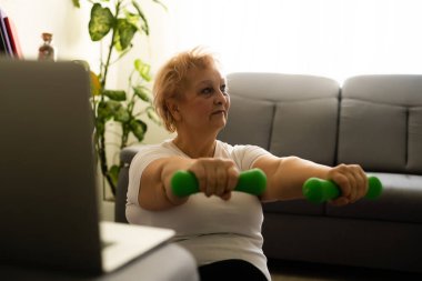 Ev sporu. Aktif kıdemli kadın dizüstü bilgisayarın önünde ısınma egzersizleri yapıyor, çevrimiçi öğretmenlerle eğitim görüyor.