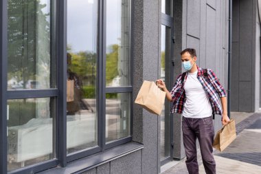 Tıbbi maskeli genç teslimatçı elinde kese kâğıdıyla yüzünde duruyor. Alışveriş çantalı hippi kurye. Güvenli çevrimiçi teslimat servisi. Coronavirus COVID-19 koruması