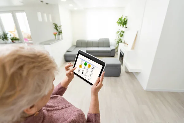 Seniorin steht zu Hause in der Küche und hält ein digitales Tablet in der Hand, das Smart-Home-System steuert — Stockfoto