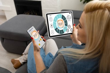 Mobil uygulamada ev sıcaklığı, güvenlik ve çevre kontrolü. Kadın elinde akıllı telefon. Arka planda oturma odası