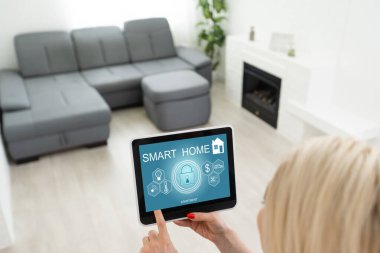 Kadın tablet üzerinde akıllı ev kontrolü uygulaması kullanıyor. Arka planda oturma odasının içi.