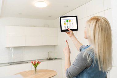 Kadın tablet üzerinde akıllı ev kontrolü uygulaması kullanıyor. Arka planda oturma odasının içi.