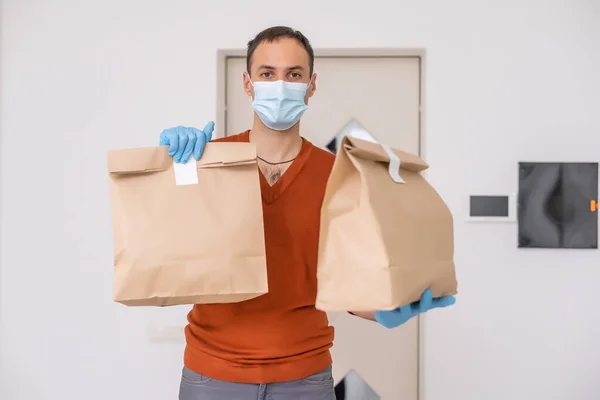 Delivery guy with protective mask and gloves holding box, bag with groceries in front of a building.