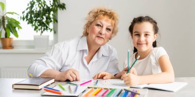 Kadın öğretmen ya da koruyucu anne, beyaz bir kız öğrencinin masada oturup ödevlerini yapmasına yardım ediyor. Çeşitli dadılar ve çocuklar evde ders çalışarak not defterinde yazmayı öğreniyorlar..