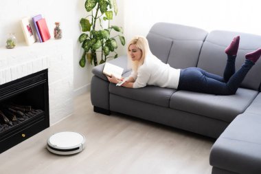 Kadın kanepede dinlenirken robot süpürge odayı temizliyor. Uzaktan kumandayla vakumu kontrol eden kadın