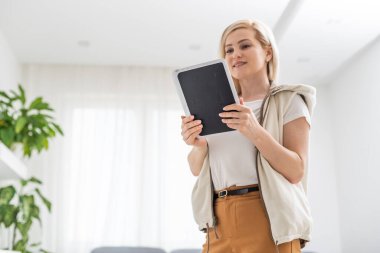Günlük giysiler içindeki çekici genç kadın dijital tablet kullanıyor, kameraya bakıyor ve evde gülümsüyor.