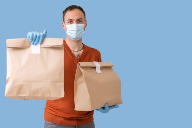 Tıbbi maskeli genç teslimatçı mavi arka planda izole edilmiş bir karton kutu taşıyor. İnternetten karantina konsepti satın al.