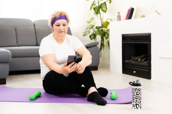 older woman training gym at home holding smartphone. Senior female workout with fitness app on the phone. Concept about senior people and sport.