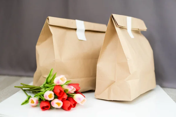 Kraft paper bags with blooming red tulips bouquet on gray background. flowers and food delivery packaging banner copy space mockup. Donations help recycling boxes