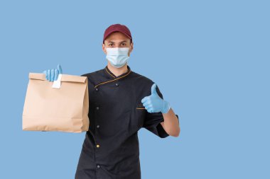 Tıbbi maskeli genç teslimatçı mavi arka planda izole edilmiş bir karton kutu taşıyor. İnternetten karantina konsepti satın al.