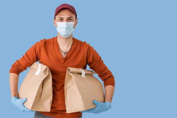 Νεαρός ντελιβεράς με ιατρική μάσκα που κρατάει και κουβαλάει ένα χαρτοκιβώτιο απομονωμένο σε μπλε φόντο. Αγοράστε τα τρόφιμα σε απευθείας σύνδεση σε καραντίνα έννοια. — Φωτογραφία Αρχείου