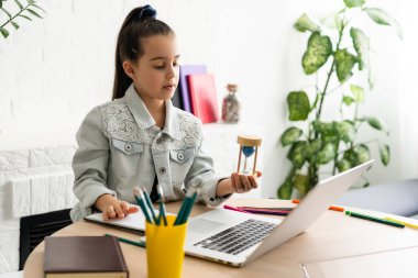 Küçük kız masada dizüstü bilgisayarla oturuyor ve elinde bir kum saati tutuyor. İnternetteki derslerin bitmesini bekliyor.