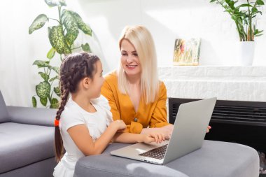 gülümseyen genç anne ve kız modern oturma odasındaki internet derslerinde ders çalışıyor.