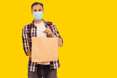 Eldiven taşıyan teslimatçı eleman, sarı arka plandaki stüdyoda izole edilmiş gıdalar içeren el işi kağıt paketi tutuyor. Servis karantinası koronavirüs virüsü 2019-ncov konsepti.