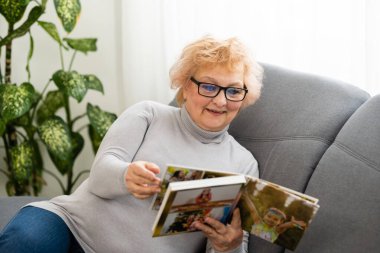 Neşeli, son sınıf öğrencisi, elinde aile fotoğrafı albümüyle evde oturmuş, yaşlı bir kadın fotobook izliyor.