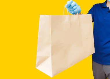 Eldiven taşıyan teslimatçı eleman, sarı arka plandaki stüdyoda izole edilmiş gıdalar içeren el işi kağıt paketi tutuyor. Servis karantinası koronavirüs virüsü 2019-ncov konsepti.