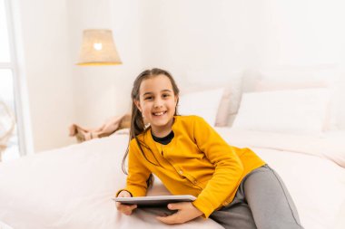 Gülümseyen küçük kız rahat koltukta uzanıyor, dijital tablet bilgisayarında online oyun oynamaktan zevk alıyor. Teknolojiye bağımlı, komik uygulamalar kullanan mutlu küçük çocuk, internet sörfü bilgisi