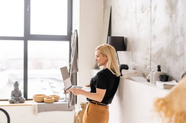 Uzaktan kumandalı ev işi, karantina sırasında serbest çalışma. Gülümseyen bayan rahat oturma odasında dizüstü bilgisayarda oturuyor ve çalışıyor.