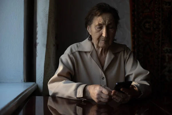 Uma avó caucasiana sênior muito velha com rugas profundas senta-se em casa, usa seu telefone inteligente na câmera frontal do braço estendido para uma chamada de vídeo. — Fotografia de Stock