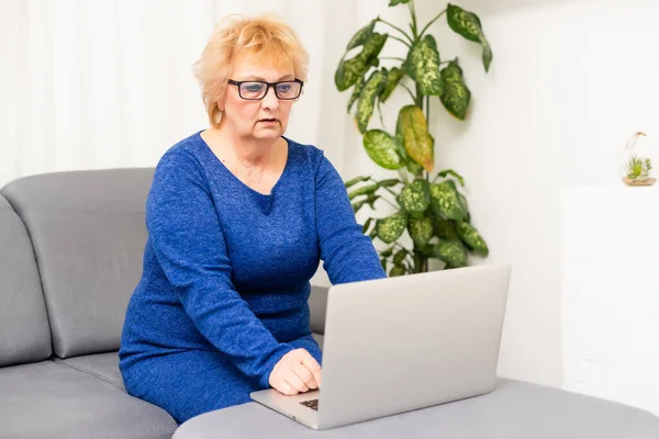 Счастливая взрослая женщина видео вызова на ноутбук работает из дома. Улыбка 60-х бизнесвумен среднего возраста разговаривает по конференции виртуальный чат с помощью компьютера в домашнем офисе сидя на диване — стоковое фото