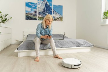 Kadın süpürgeyi uzaktan kumandayla kontrol ederken robotik süpürge odayı temizliyor..