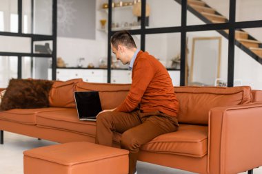 Evdeki kanepede oturan mutlu genç adam dizüstü bilgisayarda çalışıyor, gülümsüyor..