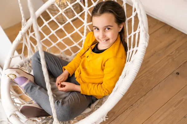 Ein schönes junges dunkelhaariges Mädchen entspannt sich in einer Schaukel. Nettes brünettes Mädchen schaukelt in einem gewebten Stuhl zu Hause. Blickt in die Kamera. — Stockfoto