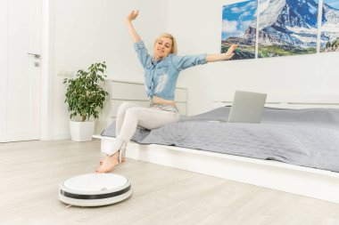 Robot süpürge oturma odasında yıkarken dizüstü bilgisayar kullanan mutlu bir kadın.