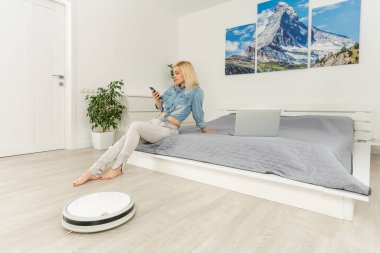 Robot süpürge oturma odasında yıkarken dizüstü bilgisayar kullanan mutlu bir kadın.