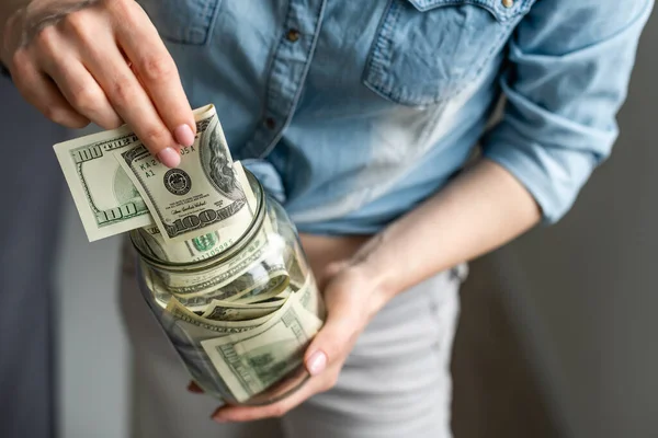 Negócio, finanças, economia, bancário e conceito de pessoas - close-up de mãos de mulher colocando-nos dinheiro dólar em frasco de pedreiro de vidro — Fotografia de Stock