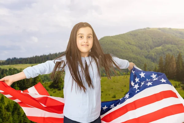 Giovane bambina che tiene bandiera americana sul cielo incredibile, montagna e prato sfondo della natura al tramonto. — Foto Stock