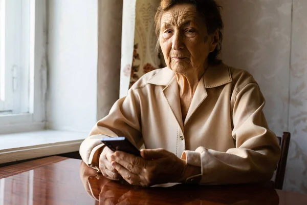 Bardzo stara, biała babcia z głębokimi zmarszczkami siedzi w domu, używa swojego smartfona na wyciągniętej przedniej kamerze ramienia do rozmowy wideo. — Zdjęcie stockowe