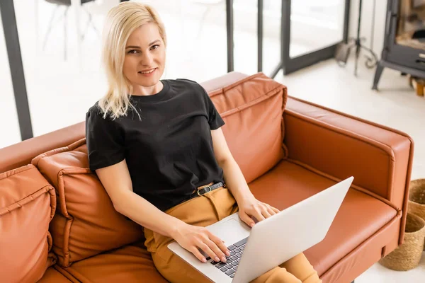 Uzaktan kumandalı ev işi, karantina sırasında serbest çalışma. Gülümseyen bayan rahat oturma odasında dizüstü bilgisayarda oturuyor ve çalışıyor.