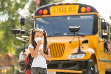 Okula dönelim. Grip virüsünden korunmak için sağlık maskesi takan kız öğrenci. Okulun arka planında bir kız öğrencinin portresi var.