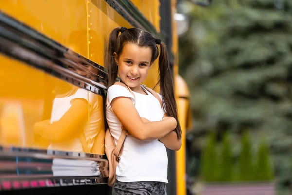 가방을 들고 버스 근처에 서 있는 귀여운 소녀 가 학교에 가는 모습을 찍기 위해 포즈를 취하고 있습니다. — 스톡 사진