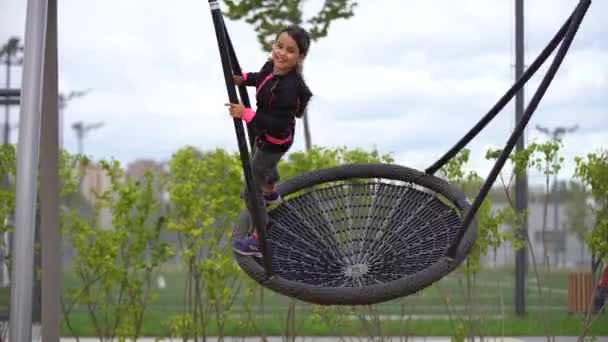 Uma menina a brincar no parque infantil. Criança feliz escalando na rede do playground, montando um balanço e bungee, estilo de vida ativo — Vídeo de Stock