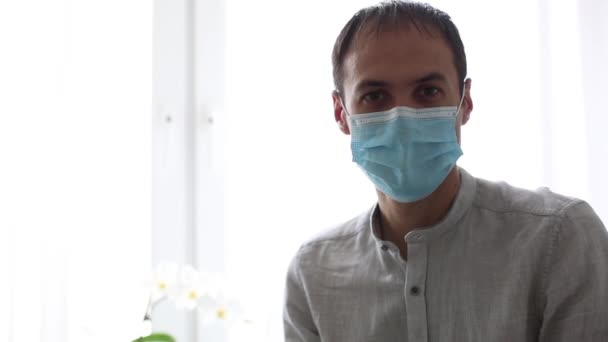 Muž v ochranné masce stojí doma na pozadí města za oknem. Koncept viru, onemocnění. — Stock video