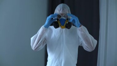 Koruyucu takım elbiseli, gözlüklü ve sağlık maskeli bir adam evdeki kanepede oturuyor ve eldiven takıyor. Koronavirüs covid-19 dünya salgınına karşı koruyucu önlemler.
