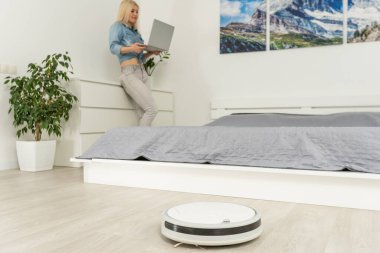 Robot süpürge oturma odasında yıkarken dizüstü bilgisayar kullanan mutlu bir kadın.
