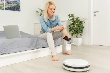 Kadın süpürgeyi uzaktan kumandayla kontrol ederken robotik süpürge odayı temizliyor..