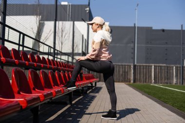 Güzel sarışın fitness kızı stadyumda esneme hareketleri yapıyor. Spor aktivitesi. Arka planda yeşil stadyum çimenleri.