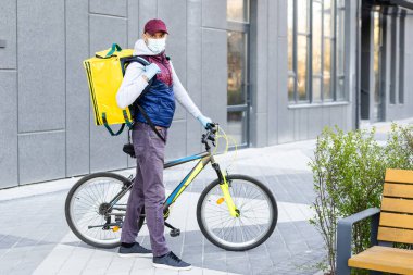 Genç kurye, şehirde bisiklet süren sarı bir termal sırt çantasıyla yiyecek getiriyor. Yemek servisi konsepti
