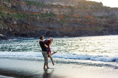 Kumsalda birlikte yürüyen tatilci çift birbirine sarılıyor. Mutlu ırklar arası genç çift, Asyalı kadın ve beyaz adam.