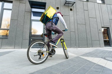 Tıbbi maskeli genç kurye, şehirde bisiklet süren sarı termal çantalı yiyecek getiriyor. Yemek servisi konsepti